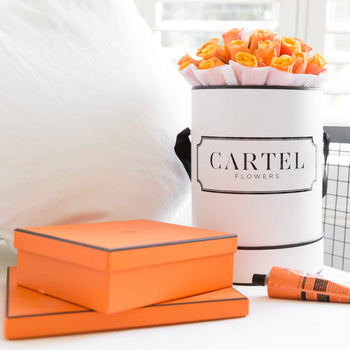 Hermes Orange Roses in a personalised monochrome white hat box with the signature Cartel Flowers branding (the worlds first personalised flowers) You can customise and personalise the writting message on the box with anything you like for any occasion. These orange roses are styled in a lifestyle photo on a cream white coloured sofa arm chair and sit next to a stack of Hermes boxes and an orange Aesop hand cream. 