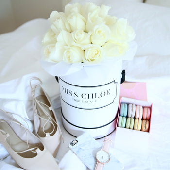 Beautiful large fresh white roses in full bloom in a personalised white hat box with a custom personalised message reading "Miss Chloe With Love" by Cartel Flowers (the worlds first personalised flowers) You can customise & personalise the writting message on the box with anything, for any occasion. Styled in a lifestyle photo on a bed along side a beige & rose gold watch, iPhone with marble phone case, a pair of beige high heels & a box of Laduree Macaroons in various flavours & colours.